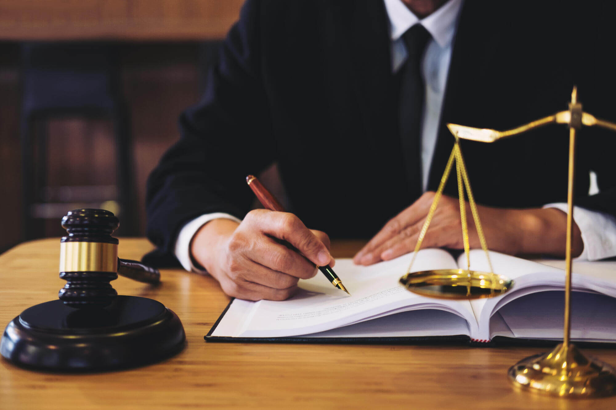 A person in a suit sits at a desk, writing in a book with a pen. A gavel and golden scales of justice on the desk symbolize law and justice, reflecting the dedication of a civil litigation attorney Florida to upholding legal standards.