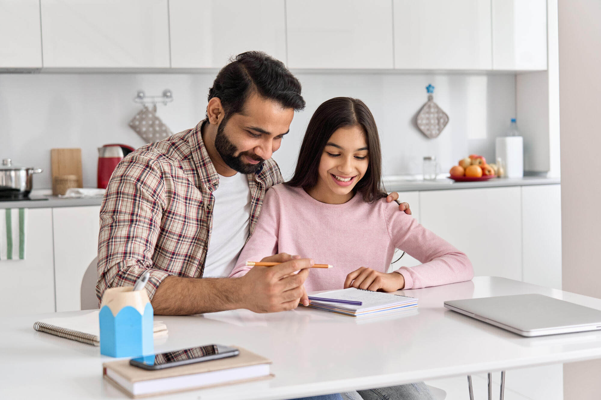 A man and a young girl sit at a kitchen table smiling as the man, a Florida attorney, helps the girl with her homework. Books, a phone, and a laptop are on the table, and the kitchen is bright and modern.
