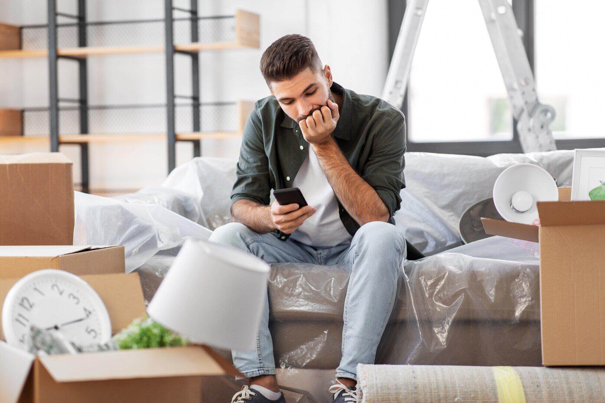 A man sits on a plastic-covered couch surrounded by moving boxes and household items, looking concerned while checking his phone—perhaps considering calling a Florida attorney for guidance during this stressful move.