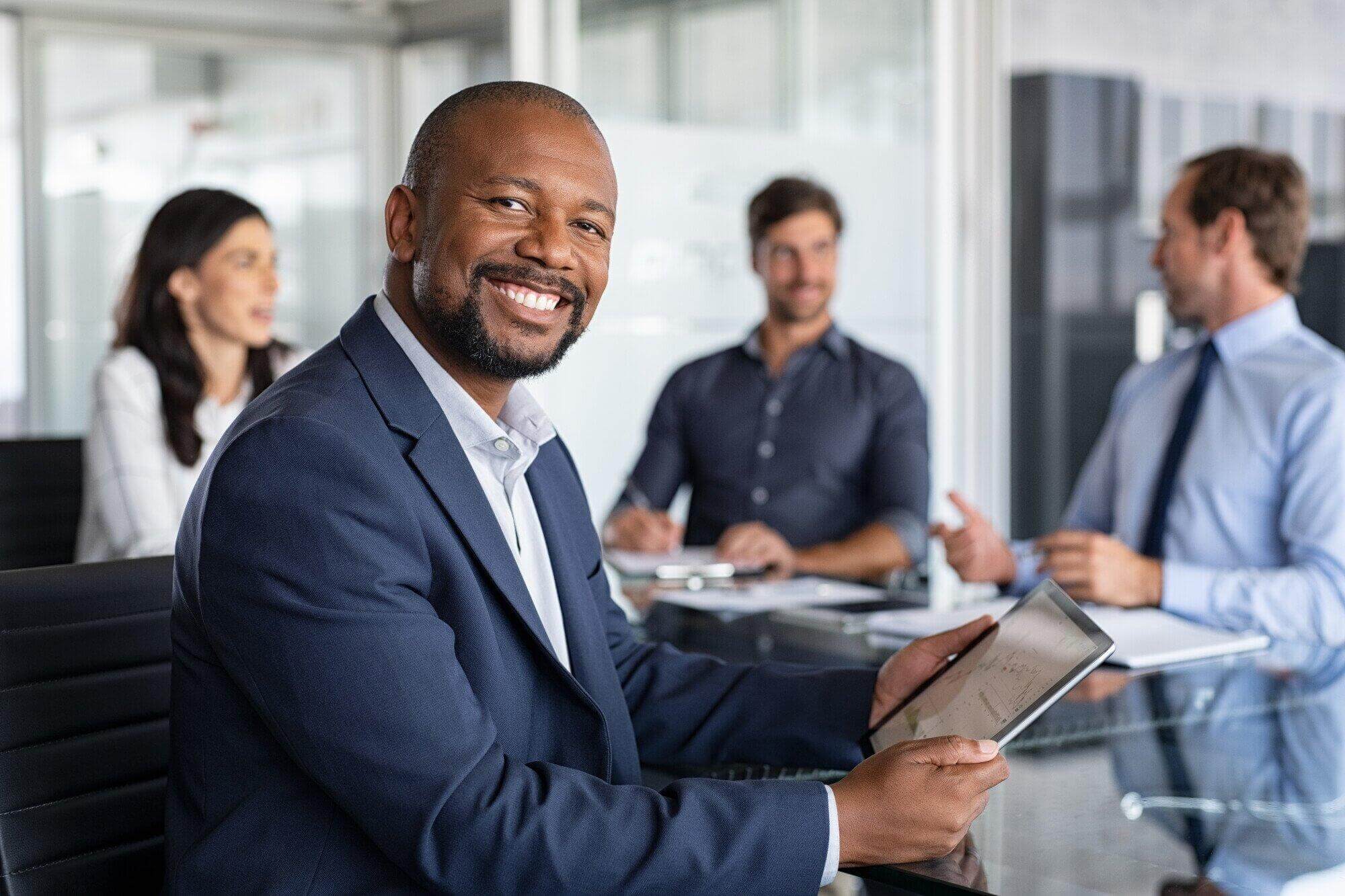 A man in a suit smiles at the camera while holding a tablet during a business meeting. Three colleagues, including a skilled civil litigation attorney Florida, sit around a conference table in a bright, modern office.