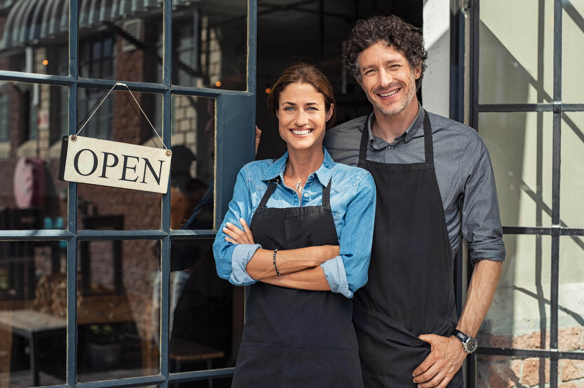 Two smiling people in aprons stand in the doorway of a shop with a glass window. An OPEN sign hangs on the door, welcoming customers—just like civil litigation attorneys in Florida welcome clients seeking resolution. Sunlight brightens the inviting entrance.