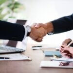 Two people in business attire shake hands across a desk with a laptop and documents, suggesting a successful meeting or agreement—an image reflecting the professionalism of Dispute Resolution Attorneys.