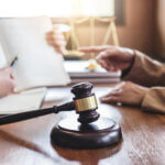 A wooden gavel rests on a desk in the foreground, while two people discuss in the background—one holding an open book and the other pointing. Legal scales are visible, suggesting the expertise of Dispute Resolution Attorneys.