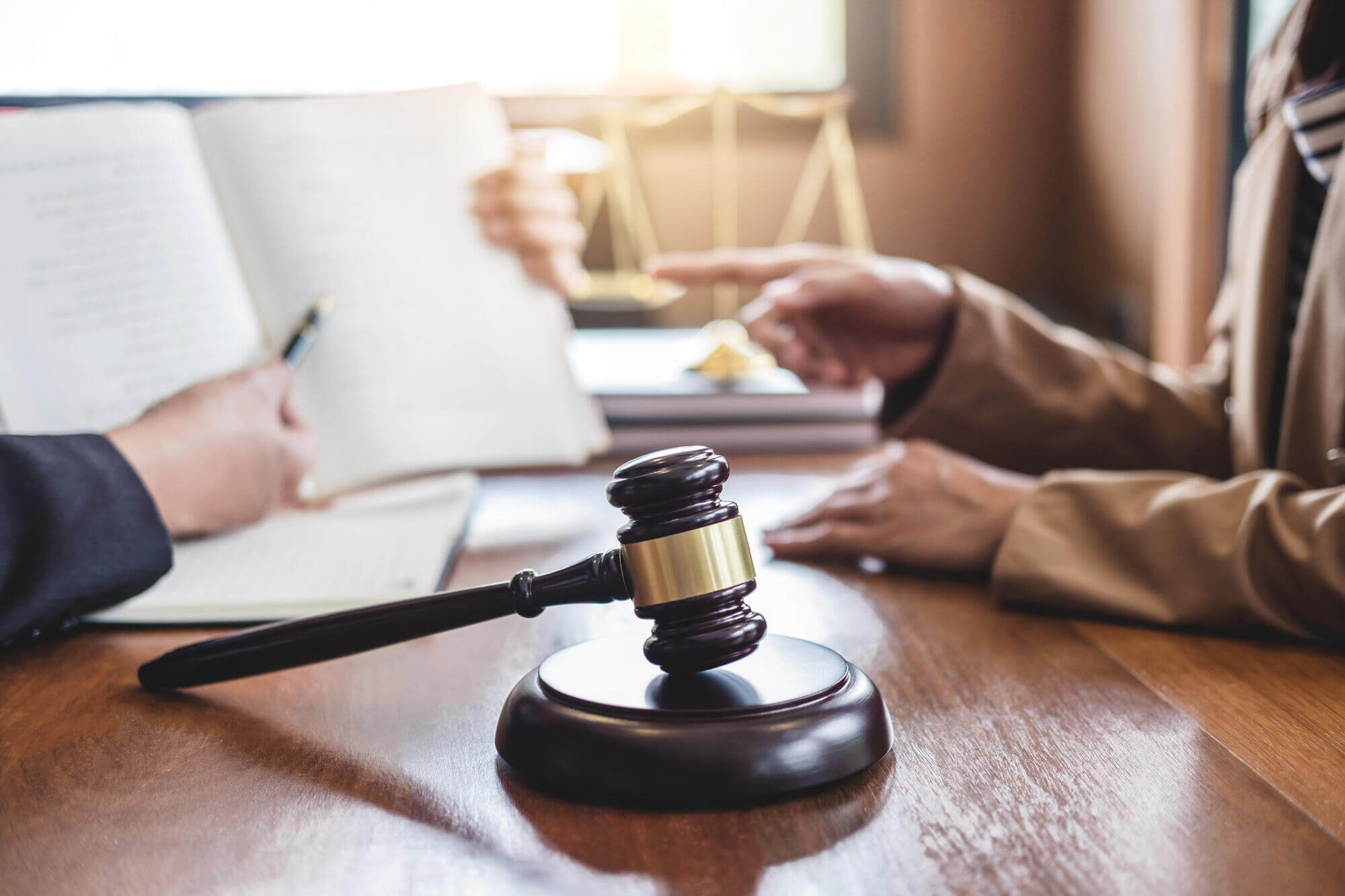 A wooden gavel rests on a desk in the foreground, while two people discuss in the background—one holding an open book and the other pointing. Legal scales are visible, suggesting the expertise of Dispute Resolution Attorneys.