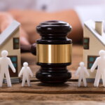 A wooden judge’s gavel is positioned in the center of a table. Around it are white paper cutout figures symbolizing family members, including adults and children. Small model houses appear on both sides in the background. The arrangement visually represents family law matters such as divorce, custody, property division, or domestic disputes.