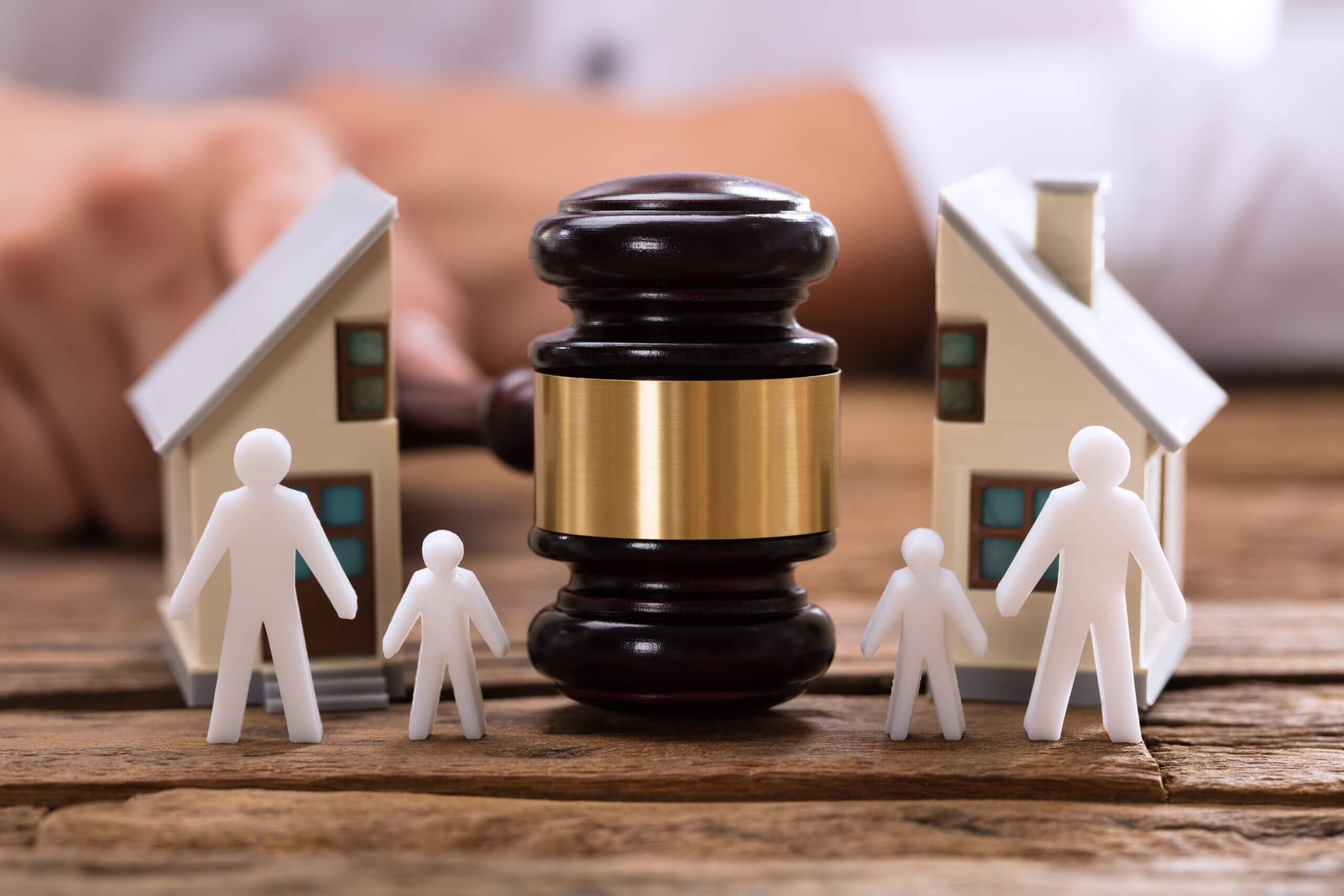 A wooden judge’s gavel is positioned in the center of a table. Around it are white paper cutout figures symbolizing family members, including adults and children. Small model houses appear on both sides in the background. The arrangement visually represents family law matters such as divorce, custody, property division, or domestic disputes.