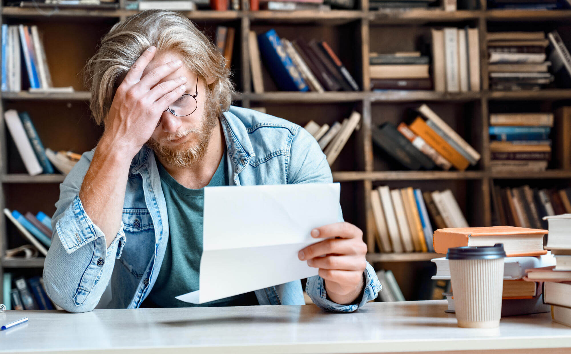A man with glasses and long hair sits at a desk, holding his head with one hand and reading a piece of paper with a concerned expression—perhaps he's seeking advice from a family law attorney. Bookshelves and a coffee cup are in the background.
